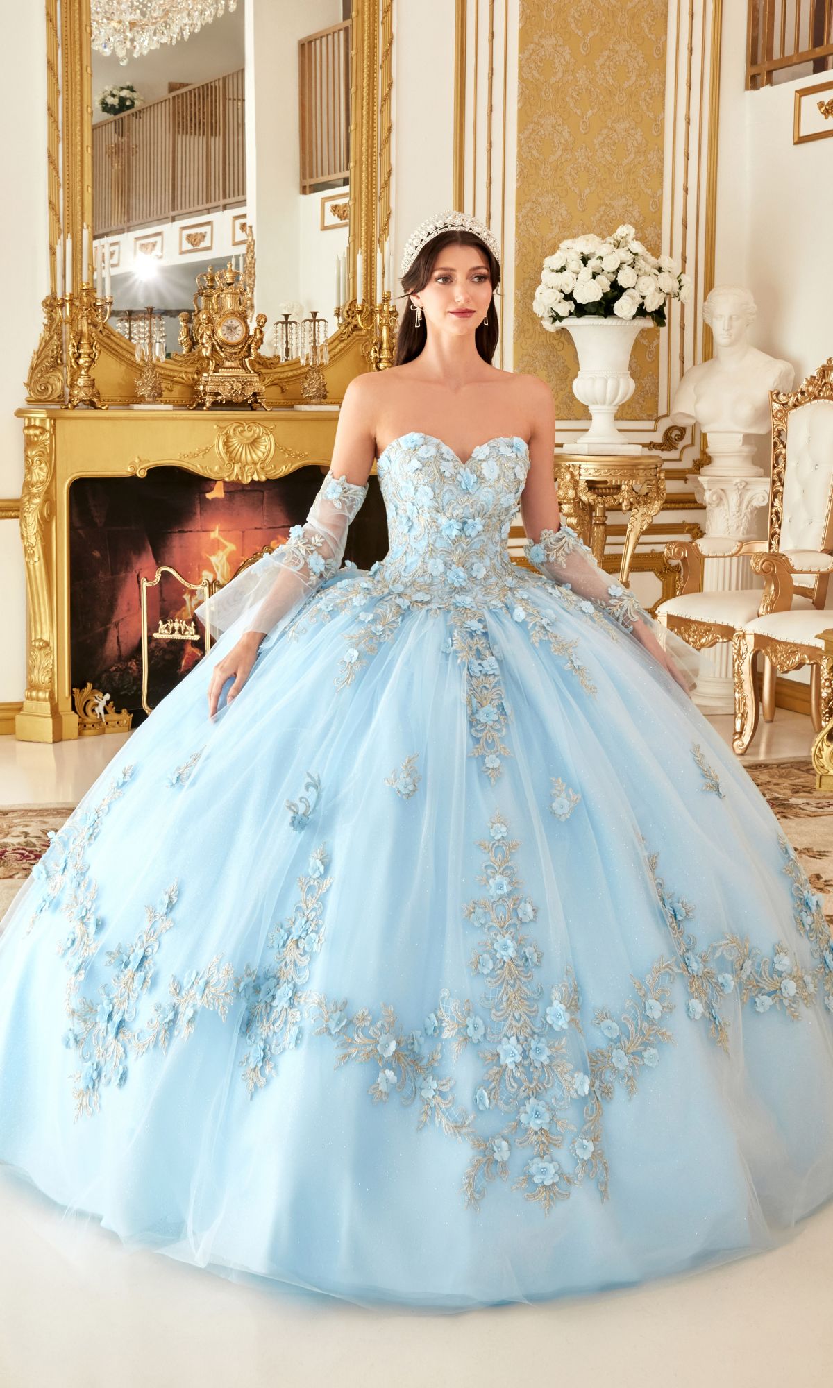 A woman in the Quinceanera Dress 15714 by Ladivine, featuring lace embroidery and a tiara, stands in an ornate room with gold accents and a lit fireplace.